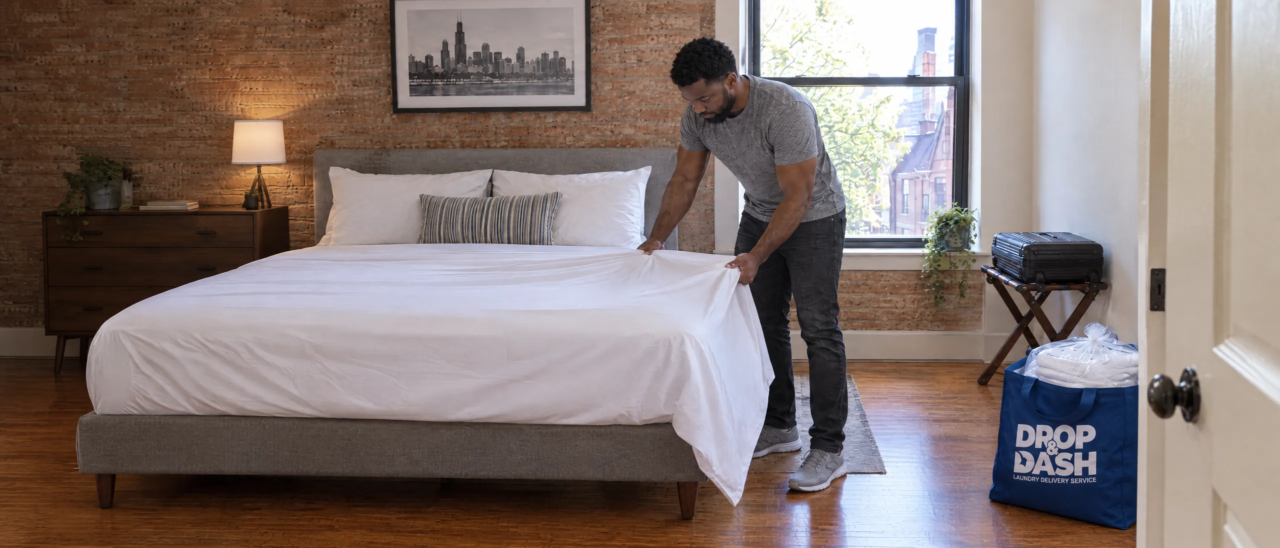 Vacation rental host making a bed with fresh white linens beside a blue Drop & Dash bag