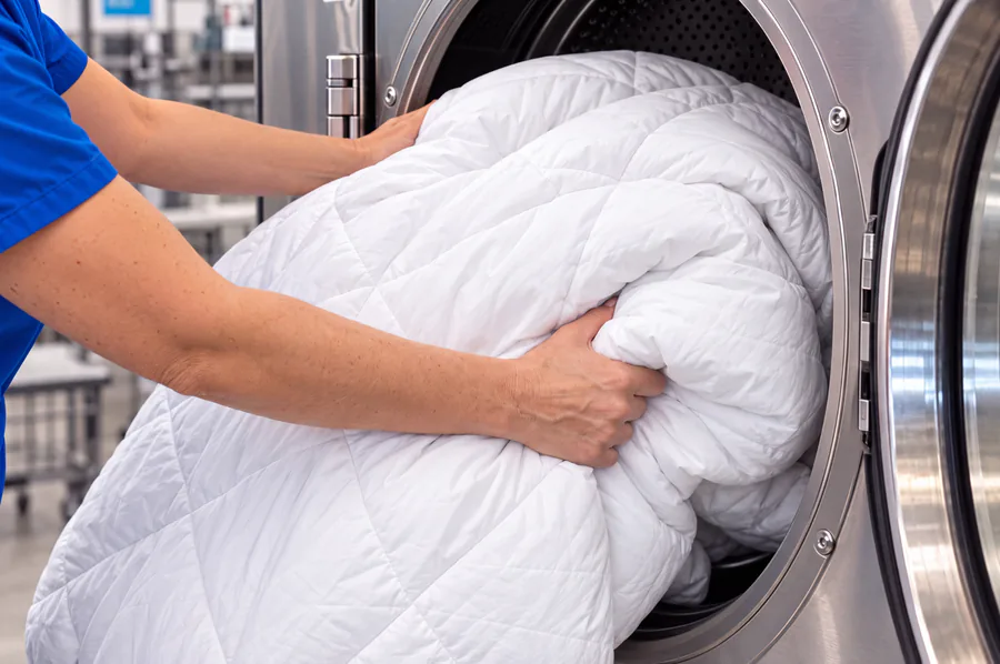 A white quilted comforter being loaded into a large commercial washing machine